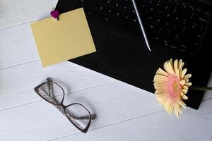 zwart laptop toetsenbord, geel leeg papier, eyewear en beige gerbera Aan een wit houten werk bureau. vrouw werkplek. foto