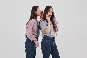 delen met geheimen. twee zussen tweeling staan en poseren in de studio met witte achtergrond foto