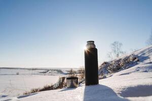 een zwart thermosfles van thee staat Aan de sneeuw achter de zon. buitenshuis recreatie gedurende een winter wandeltocht, een mok met mosterd thee in natuur. foto