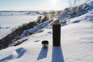 de concept van een thermosfles tegen de achtergrond van winter natuur. een zwart thermosfles met een mok staat in de sneeuw. zonlicht, schaduw van de object, toerist apparatuur. foto