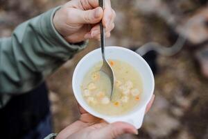 een kom van soep is schot detailopname, voedsel Aan een wandeltocht, toerist ontbijt in natuur, aardappel soep is gegeten met een lepel, heet voedsel, een wit bord, een meisjes handen. foto