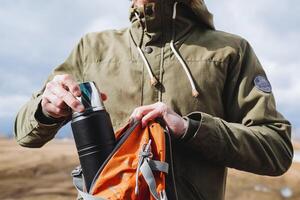 een toerist duurt uit van een rugzak een zwart metaal thermosfles, camping apparatuur, een vacuüm thermosfles voor heet drankjes, uitrusting Aan een wandeltocht, gerechten van een toerist. foto