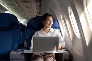 geslaagd jong Aziatisch bedrijf vrouw zit in vliegtuig cabine vlak en werken van overal. vliegend Bij eerste klasse foto