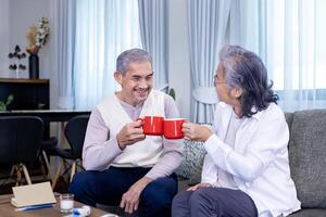 paar van gezond Aziatisch senior vader en moeder zittend Aan bankstel met gelukkig glimlach Bij pensioen huis drinken heet thee naar vieren hun vakantie samen voor ouderling zorg en uitgeven waardevol tijd foto