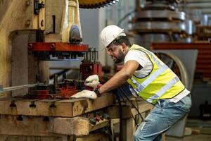 Afrikaanse Amerikaans industrieel arbeider is gebruik makend van hydraulisch macht druk op machine naar maken metaal en staal een deel terwijl werken binnen de metaal vel verzinkt dak fabriek voor veiligheid industrie foto