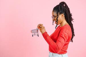 Afrikaanse Amerikaans meisje met boodschappen doen kar Aan roze achtergrond. de concept van winstgevend verkoop of aankopen foto