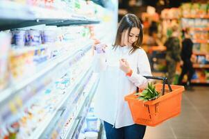 vrouw aan het doen kruidenier boodschappen doen Bij de supermarkt, ze is leunend Aan de boodschappen doen kar en Verbinden met haar telefoon, apps en kleinhandel concept foto