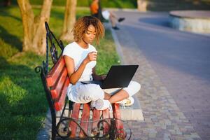 portret van Afrikaanse Amerikaans vrouw met afro kapsel zittend Aan de houten bank buitenshuis en werken Aan laptop foto