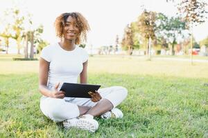 attent schattig gemengd vrouw Internationale leerling met gekruld haar- is zittend Aan vers gras met modern laptop in openbaar park, leunend Aan appel boom en weemoedig op zoek terzijde gedurende haar breken foto