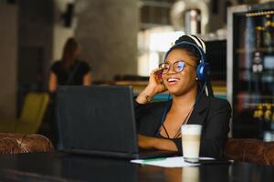 gelukkig zwart meisje in draadloze hoofdtelefoons aan het studeren online, gebruik makend van laptop en nemen notities, cafe interieur, kopiëren ruimte foto