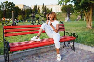 mooi jong Afrikaanse Amerikaans vrouw met afro kapsel vervelend wit jurk. mooi meisje zittend in de park met een smartphone. foto