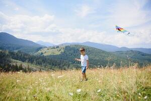 weinig jongen rennen Aan een achtergrond van bergen met vlieger. zonnig zomer dag. gelukkig kinderjaren concept. foto