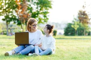 mam en dochter spelen in de laptop buitenshuis, lachend en genieten van de zomer zon Aan de groen gras in de park, familie buitenshuis in de park foto