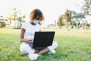 jong zwart vrouw Bij werk Aan haar laptop in keer van epidemie buitenshuis, gebruik van beschermend masker, jong leerling Bij de park foto