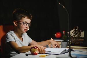 jongen leren lessen in de huis instelling Bij de tafel in de licht van een tafel lamp. foto