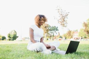 jong aantrekkelijk met een donkere huid college leerling vervelend tank top en jeans zittend Aan de gazon Bij campus Aan zonnig dag, werken Aan haar scriptie gebruik makend van laptop computer, op zoek bezig en geconcentreerd foto