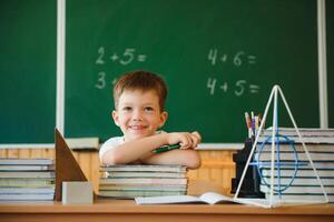 terug naar school. een schooljongen is lezing een boek terwijl zittend Bij de tafel voor de eerste tijd Bij school- Aan de achtergrond van de school- schoolbord. foto