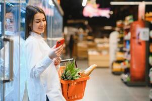 glimlachen jong vrouw gebruik makend van mobiel telefoon terwijl boodschappen doen in boodschappen doen op te slaan foto