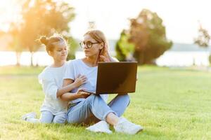 mam en dochter spelen in de laptop buitenshuis, lachend en genieten van de zomer zon Aan de groen gras in de park, familie buitenshuis in de park foto