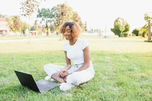 menselijk en technologie concept. charmant Afrikaanse vrouw met kort pixie kapsel genieten van zonnig het weer, zittend Aan de gazon in voorkant van laptop computer in de openbaar tuin, aan het wachten voor haar vriend foto