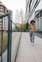 vrouw freelancer is pratend telefoon terwijl staand met laptop Aan modern gebouw achtergrond foto