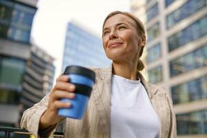 vrouw manager gaan naar werk terwijl drinken koffie Aan kantoor gebouw achtergrond en looks weg foto