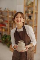 portret van jong vrouw pottenbakker in schort met mok op zoek Bij camera terwijl poseren in werkplaats foto