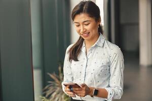 glimlachen vrouw kantoor arbeider op zoek Aan smartphone terwijl staand Aan modern coworking achtergrond foto