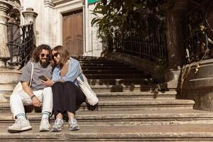 mooi hipster paar in liefde zittend trap Aan oud stad straat en op zoek Aan telefoon foto