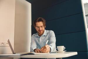 glimlachen mannetje freelancer maken aantekeningen gedurende werken Aan laptop sitiing bureau in coworking foto