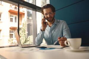mannetje baas pratend telefoon en maken aantekeningen terwijl zittend in modern kantoor en werken Aan laptop foto