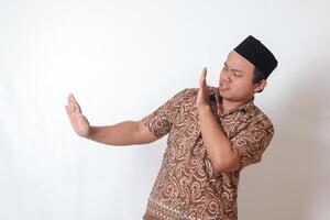 portret van onaangenaam Aziatisch Mens vervelend batik overhemd en songkok vormen een hand- gebaar naar vermijden iets. reclame concept. geïsoleerd beeld Aan grijs achtergrond foto