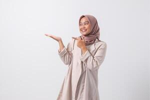 portret van opgewonden Aziatisch hijab vrouw in gewoontjes pak richten en tonen Product in haar kant met vinger. zakenvrouw concept. geïsoleerd beeld Aan wit achtergrond foto