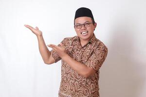 portret van opgewonden Aziatisch Mens vervelend batik overhemd en songkok glimlachen en op zoek Bij de camera richten met twee handen en vingers naar de kant. geïsoleerd beeld Aan grijs achtergrond foto