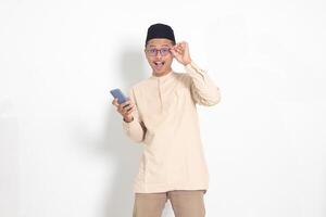 portret van verrast Aziatisch moslim Mens in koko overhemd met kalotje Holding mobiel telefoon, tonen Wauw geschokt gezicht uitdrukking. reclame en sociaal media concept. geïsoleerd beeld Aan wit achtergrond foto