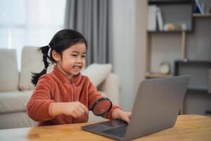 opgewonden jong meisje ontdekken met vergroten glas en laptop. gelukkig Aziatisch meisje gebruik makend van een vergroten glas naar onderzoeken Aan een laptop, aan het leren met vreugde Bij huis. onderwijs voor kind leerling concept. foto