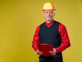 portret van een senior ingenieur met map in handen en helm op zoek naar de camera. geïsoleerd Aan geel achtergrond foto