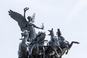 bronzen beeldhouwwerk van een gevleugeld vrouw figuur met een laurier lauwerkrans, leidend een wagen getrokken door vier paarden tegen een Doorzichtig lucht. foto