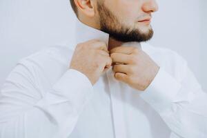 een Mens in een wit overhemd staat door de venster in de kamer en vastmaakt de toetsen Aan zijn halsband en mouwen. kijk maar Aan hand. elegant bedrijf portret van een Mens, detailopname foto. de bruidegom is voorbereidingen treffen. foto