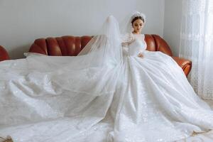 een ongelooflijk mooi bruid zit Aan een bed in haar slaapkamer. bruid ochtend. een prachtig bruid in een mooi jurk is zittend Aan een bed in een helder kamer . foto