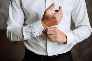 een Mens in een wit overhemd staat door de venster in de kamer en vastmaakt de toetsen Aan zijn halsband en mouwen. kijk maar Aan hand. elegant bedrijf portret van een Mens, detailopname foto. de bruidegom is voorbereidingen treffen. foto