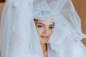 portret van een ongelooflijk mooi meisje bruid in een wit gewaad in de slaapkamer, de bruid poses Holding haar sluier in haar handen en covers haarzelf met het. foto