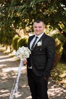 portret van een knap jong bruidegom Aan een herfst dag buitenshuis in een elegant pak en Holding een bruiloft boeket van bloemen. buitenshuis foto. knap Mens in een bedrijf pak. foto