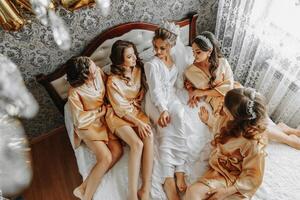 jong bruidsmeisjes in zijde Jurken in een hotel kamer. mooi Dames vieren een vrijgezellin partij zittend Aan de bed in themed pyjama. foto