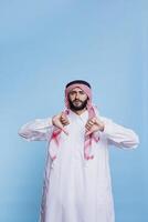 ontevredenheid Arabisch vervelend traditioneel kleren poseren met duimen naar beneden en op zoek Bij camera met echt uitdrukking. moslim Mens gekleed in thobe en ghutra en tonen afkeuring gebaar foto