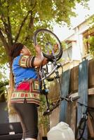actief sportief Afrikaanse Amerikaans dame waarborgt fiets wiel is beveiligen voor zomer buitenshuis vrije tijd wielersport. gezond zwart vrouw fietser veilig opnieuw bevestigen fiets band in huis tuin. foto