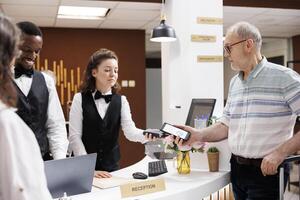 ouderen paar cheques in Bij hotel ontvangst, bijgestaan door vriendelijk multi-etnisch personeel. Kaukasisch oud Mens Bij voorkant bureau maken betaling Aan pos terminal met smartphone voor reservering en boeking. foto