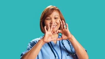 dichtbij omhoog schot van blij gekwalificeerd bipoc verpleegster aan het doen hart symbool vorm met handen. kliniek werknemer tonen liefde gebaren terwijl Bij werk, geïsoleerd over- blauw studio achtergrond foto