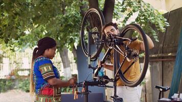 enthousiast Kaukasisch vriendje onderzoeken wiel en pedalen terwijl vriendin haalt op deskundige werk gereedschap voor repareren fiets. multiraciaal paar buitenshuis pratend en werken Aan beschadigd fiets. foto