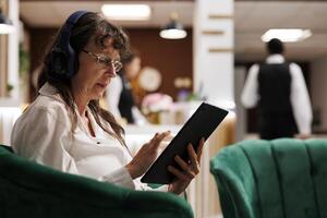 gedetailleerd schot van senior vrouw ontspannende met hoofdtelefoons en surfing de netto Aan tablet in hotel ontvangst. dichtbij omhoog van ouderen vrouw toerist Aan bank, luisteren naar muziek- en browsen de internetten. foto